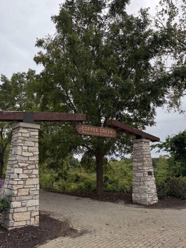 Stone entrance arch with a Coffee Creek Watershed Preserve sign surrounded by trees and greenery.